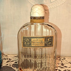 May include: A vintage clear glass perfume bottle with a textured surface and a white, textured stopper. The label reads "Eau de Cologne, Grenoville". The bottle is empty and sits on a lace doily.