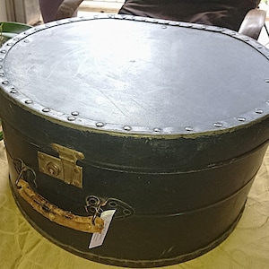Op de afbeelding: Een vintage zwarte hoedendoos met een afgeronde bovenkant en metalen accenten. De doos heeft een messing sluiting en een handvat. Een lichtgekleurd houten voorwerp staat naast de hoedendoos. De hoedendoos staat op een tafel.