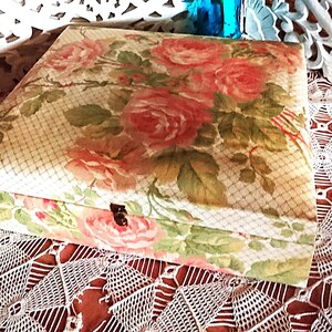 Op de afbeelding: Een rechthoekige doos met een bloemenrozenpatroon in roze en groen. De doos is gesloten met een kleine zwarte sluiting. Twee turquoise flessen en witte bloemen staan op de achtergrond.
