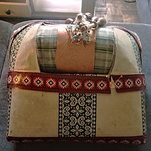 Puede incluir: Una caja de costura rectangular con una parte superior a cuadros y laterales de tela estampada. La caja está adornada con una banda decorativa con un diseño geométrico en rojo y negro. Se adjuntan alfileres y elementos decorativos en la parte superior.