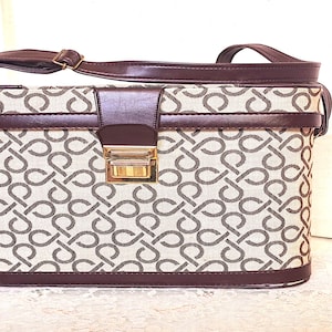 May include: A rectangular structured handbag featuring a beige fabric body with a repeating dark brown geometric pattern. The bag includes dark brown trim, a matching adjustable shoulder strap, and a central gold-tone metal clasp closure.