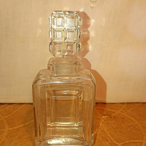 May include: A clear, vintage perfume bottle with a square base and a faceted, geometric stopper. The bottle is made of glass and has a classic, elegant design. The bottle is empty and ready to be filled.