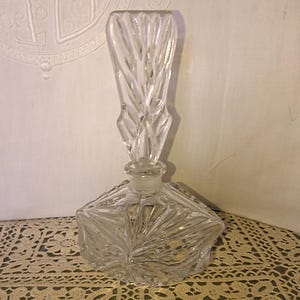 May include: A clear, vintage perfume bottle with a geometric design. The bottle has a square base and a tall, decorative stopper. The glass is intricately cut with a starburst pattern on the base and vertical lines on the stopper.
