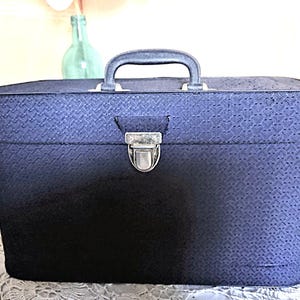 May include: A navy blue rectangular box with a handle and a silver clasp. The box has a textured pattern and a handle on top. The clasp is centered on the front. The box is sitting on a white lace surface.