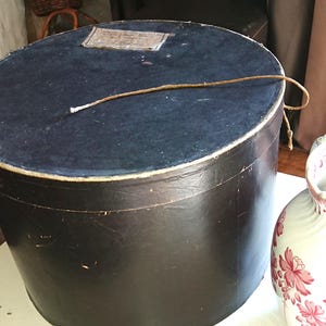 Op de afbeelding: Een grote, ronde, zwarte hoedendoos met een donkerblauwe bovenkant en een label. Een decoratieve witte en roze bloemenkan staat naast de hoedendoos. De hoedendoos heeft een touwtje eromheen.