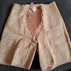 May include: Beige high-waisted shapewear shorts with a floral lace pattern. The shorts have a satin panel in the front and lace trim along the leg openings. The garment is designed to smooth and shape the midsection.