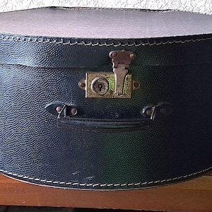 Op de afbeelding: Een vintage zwarte hoedendoos met een getextureerde buitenkant en een afgeronde bovenkant. De doos heeft een metalen sluiting en handvat, met witte stiksels langs de randen. De doos staat op een houten oppervlak.