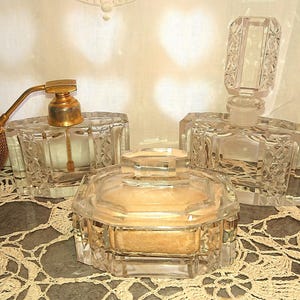 May include: A collection of vintage crystal perfume bottles and a powder jar. The bottles feature detailed cut glass patterns and gold-coloured accents. One bottle has a bulb atomizer. The items are displayed on a lace doily.