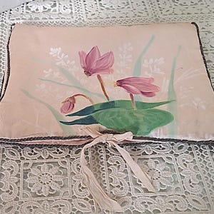May include: A rectangular, blush-colored pillow with a floral design. The artwork features pink flowers, green leaves, and white accents. The pillow has a dark gray border and a cream-colored ribbon tied in a bow.