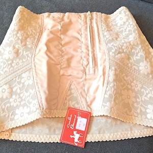 May include: A vintage peach-colored corset with floral lace detailing. The corset features a front zipper, a satin panel, and a scalloped hem. A red tag with the brand name "Caroline Bernard" is attached.