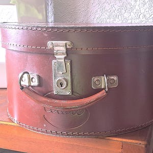 Op de afbeelding: Een vintage bruinleren hoedenkist met een afgeronde vorm. De kist heeft een metalen sluiting, handvat en decoratieve stiksels. De handgreep is van gebogen, bruin leer. De kist staat op een houten oppervlak.