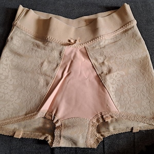 May include: Beige shapewear shorts with a high waistband and a decorative bow. The shorts feature a floral lace pattern on the front and sides, with a smooth, pink panel in the center. The edges are trimmed with delicate lace.