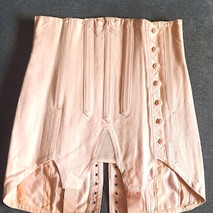 May include: A vintage, pale pink corset with vertical seams and decorative stitching. The corset features a row of buttons on one side and eyelets for lacing on the other. It has a decorative embroidered detail at the top and suspender attachments.