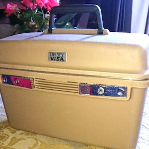 May include: A tan Delsey Visa travel case with a black handle and silver hardware. The case features two colourful latches and a Delsey Visa logo. The rectangular case has a slightly textured surface.