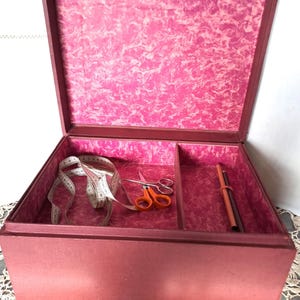 Op de afbeelding: Open bordeauxkleurige naaidoos met een roze gemarmerd interieur. Binnenin bevinden zich een witte meetlint, een oranje schaar en twee potloden. De doos staat op een met kant bedekt oppervlak.