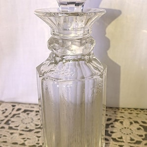 May include: A clear, faceted glass decanter with a matching stopper. The stopper has a rounded, geometric design. The decanter's body is rectangular with vertical grooves. The design is elegant and classic.