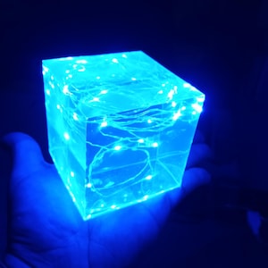 May include: A clear cube with a blue glow from LED lights inside. The cube is being held in a hand.