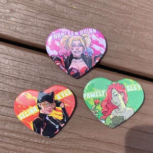 May include: Three heart-shaped pins featuring characters from the DC Comics universe. The first pin features Harley Quinn with the text "Harley Quinn". The second pin features Catwoman with the text "Selina Kyle". The third pin features Poison Ivy with the text "Pamela Isley".