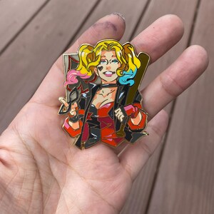 May include: Gold-coloured pin featuring a cartoon illustration of Harley Quinn. She is wearing a red and black costume, holding a baseball bat and a pair of glasses. Her hair is styled in pigtails with pink and blue streaks.