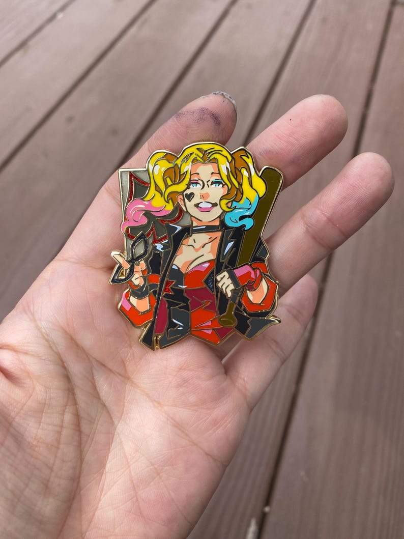 May include: Gold-coloured pin featuring a cartoon illustration of Harley Quinn. She is wearing a red and black costume, holding a baseball bat and a pair of glasses. Her hair is styled in pigtails with pink and blue streaks.