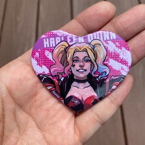 May include: A heart-shaped button with a cartoon image of Harley Quinn. The button has a pink background with white and black accents. The text "Harley Quinn" is printed in white letters on the button.