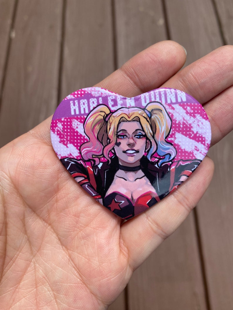 May include: A heart-shaped button with a cartoon image of Harley Quinn. The button has a pink background with white and black accents. The text "Harley Quinn" is printed in white letters on the button.