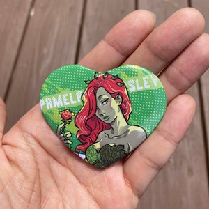 May include: A heart-shaped button with a green background and white polka dots. A cartoon image of Poison Ivy holding a red rose is in the center. The text "Pamela Isley" is on the left and "Sley" is on the right.