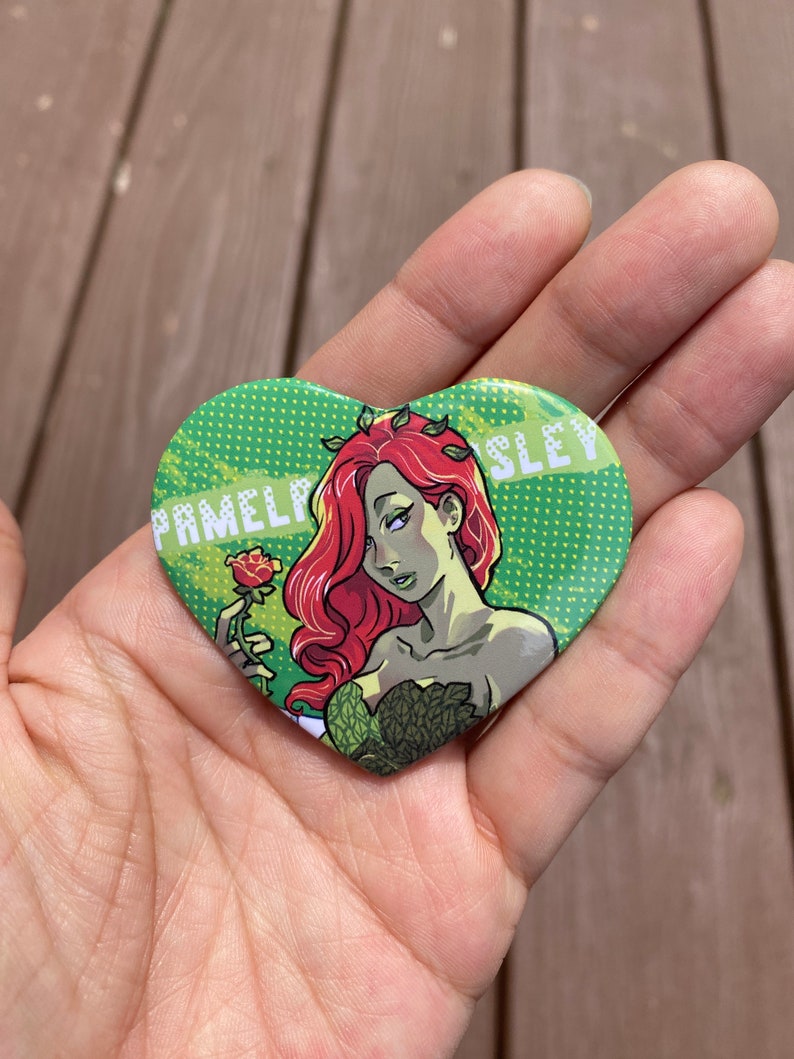 May include: A heart-shaped button with a green background and white polka dots. A cartoon image of Poison Ivy holding a red rose is in the center. The text "Pamela Isley" is on the left and "Sley" is on the right.