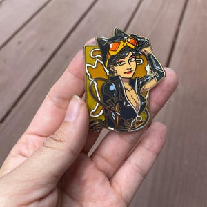 May include: Gold-tone enamel pin featuring a cartoon illustration of Catwoman in a black leather suit with a yellow cat symbol on her chest. She is wearing a black mask and goggles, and has her arm raised in a salute.