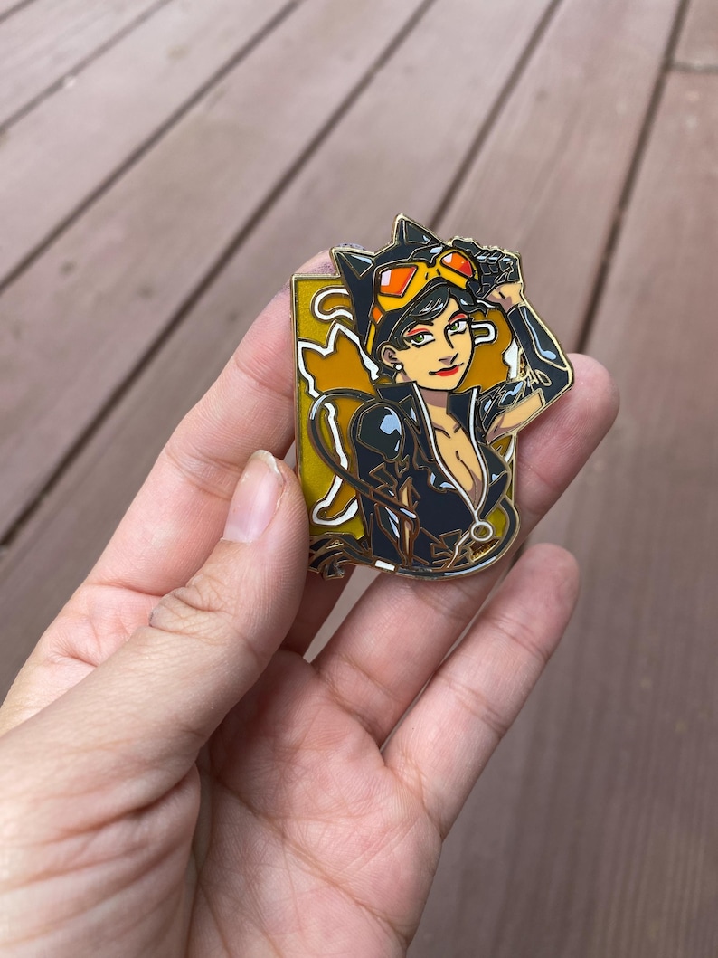 May include: Gold-tone enamel pin featuring a cartoon illustration of Catwoman in a black leather suit with a yellow cat symbol on her chest. She is wearing a black mask and goggles, and has her arm raised in a salute.