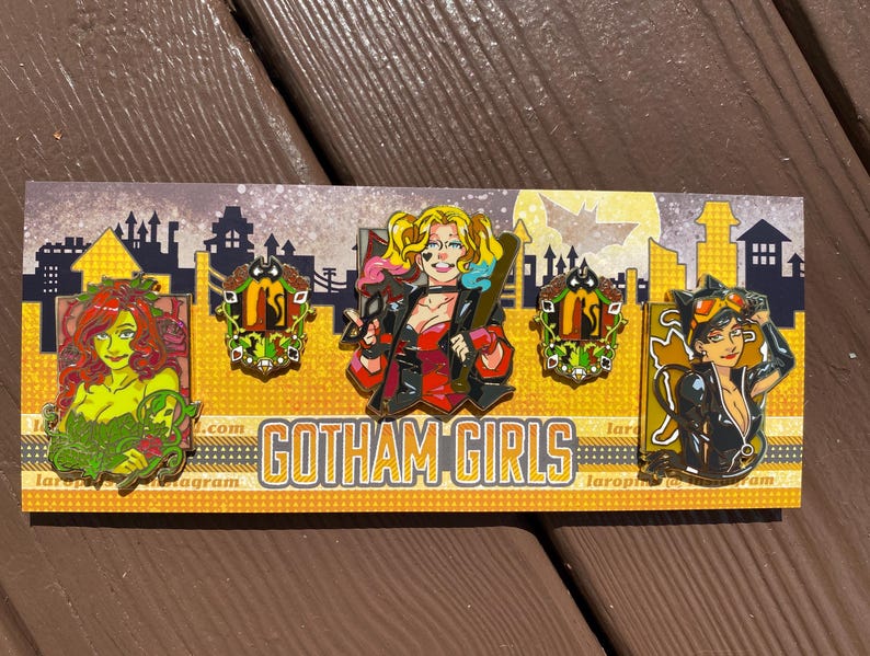 May include: Four enamel pins featuring characters from the DC Comics universe. The pins are on a yellow background with the text "Gotham Girls" in black. The characters are Poison Ivy, Harley Quinn, Catwoman, and a Gotham City crest.