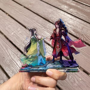 May include: A clear acrylic stand featuring two anime-style figures holding hands. One figure wears a green and yellow outfit and holds a fan. The other figure is in a red and black outfit. The base of the stand has text.