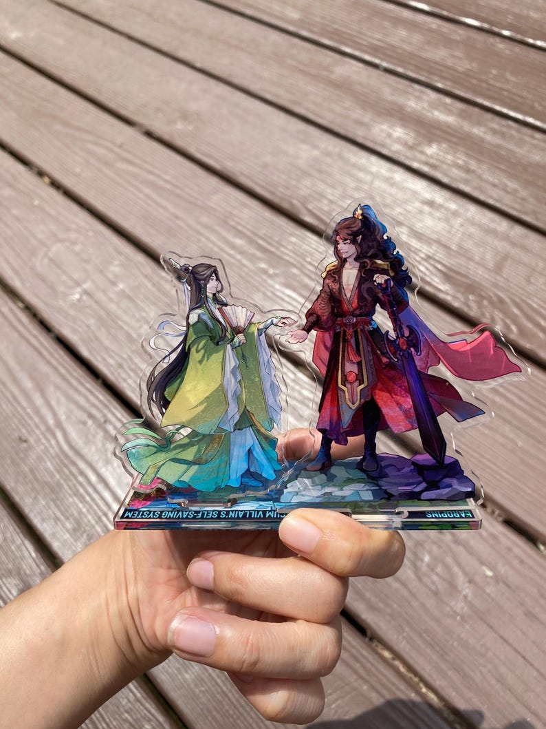 May include: A clear acrylic stand featuring two anime-style figures holding hands. One figure wears a green and yellow outfit and holds a fan. The other figure is in a red and black outfit. The base of the stand has text.