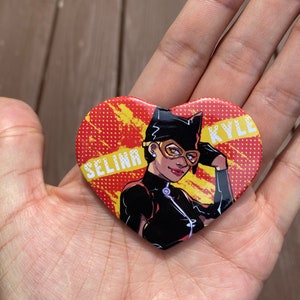 May include: A heart-shaped button with a red and yellow background. The button features a cartoon image of a woman wearing a black catsuit and a black mask. The text "SELINA KYLE" is printed on the button.