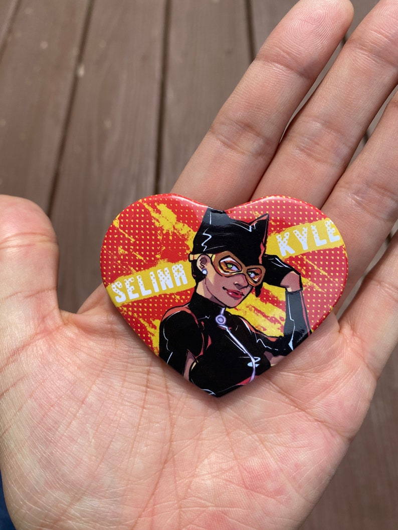 May include: A heart-shaped button with a red and yellow background. The button features a cartoon image of a woman wearing a black catsuit and a black mask. The text "SELINA KYLE" is printed on the button.