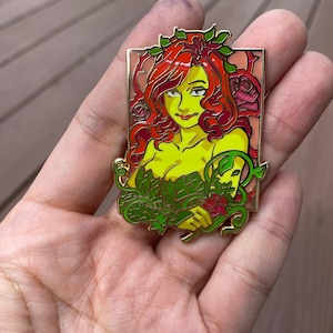 May include: A gold-coloured enamel pin featuring a cartoon depiction of Poison Ivy, a DC Comics supervillain. She has red hair, green leaves, and a red rose in her hand. The pin is set against a pink and green background.