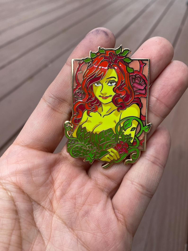 May include: A gold-coloured enamel pin featuring a cartoon depiction of Poison Ivy, a DC Comics supervillain. She has red hair, green leaves, and a red rose in her hand. The pin is set against a pink and green background.