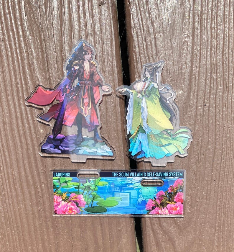 May include: Three acrylic character standees featuring anime-style illustrations. One figure wears red and black robes, another wears green and blue robes. A rectangular standee displays the text "THE SCUM VILLAIN'S SELF-SAVING SYSTEM" with floral and water lily graphics.