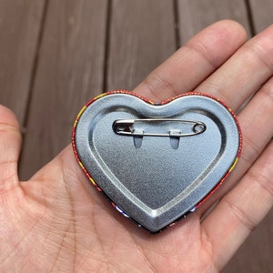 May include: The back of a heart-shaped button with a safety pin attached. The button is silver with a red and yellow patterned border.