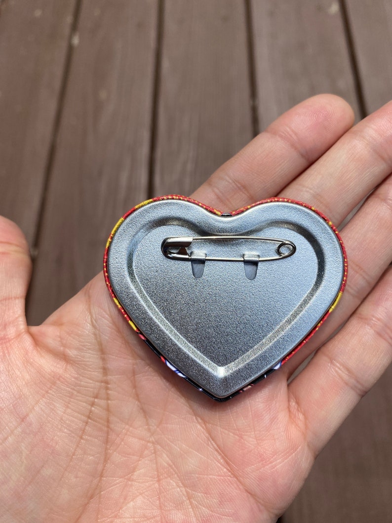 May include: The back of a heart-shaped button with a safety pin attached. The button is silver with a red and yellow patterned border.