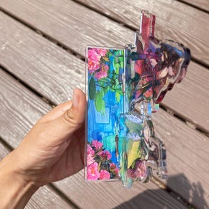 May include: A clear acrylic decorative piece featuring a floral and landscape design. The piece has a layered effect with pink flowers, blue water, and green foliage. The edges are clear, and the piece is held by a hand.