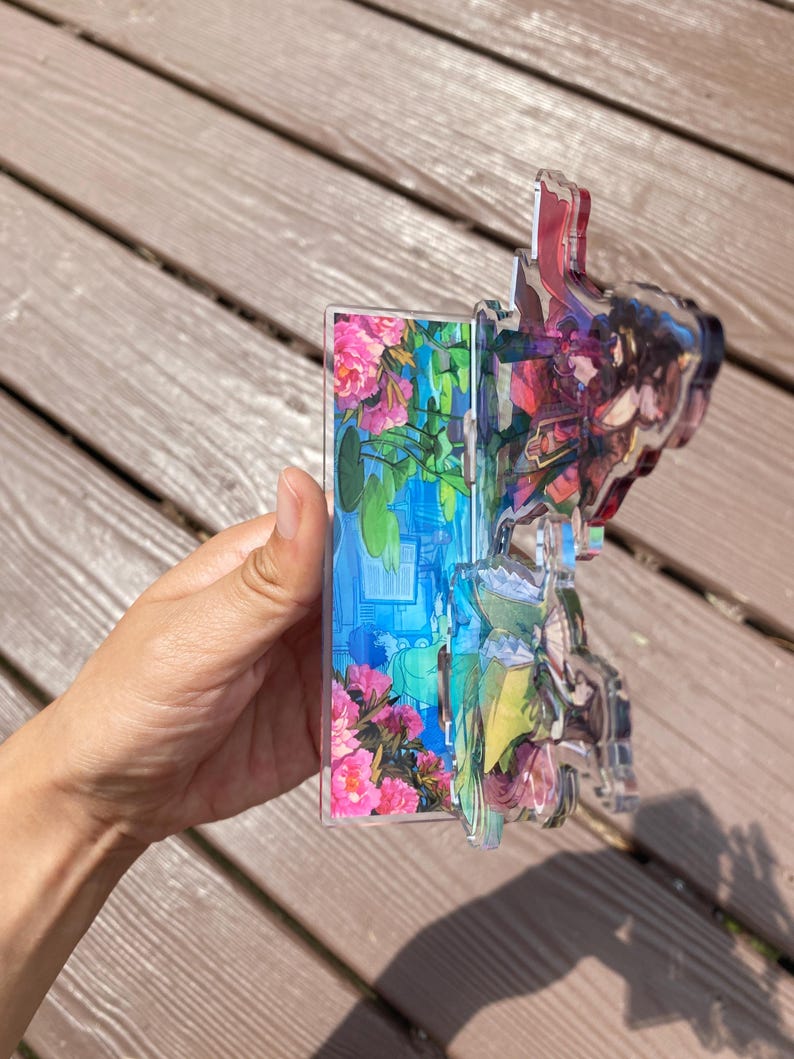 May include: A clear acrylic decorative piece featuring a floral and landscape design. The piece has a layered effect with pink flowers, blue water, and green foliage. The edges are clear, and the piece is held by a hand.