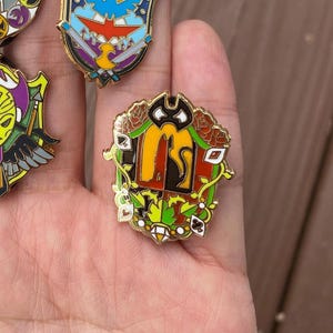 May include: A gold-coloured enamel pin featuring a black cat, red, orange, and green leaves, and playing card symbols. The pin is shaped like a shield.