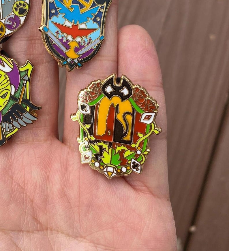 May include: A gold-coloured enamel pin featuring a black cat, red, orange, and green leaves, and playing card symbols. The pin is shaped like a shield.
