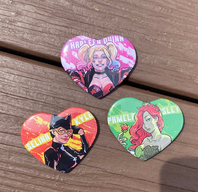 May include: Three heart-shaped pins featuring characters from the DC Comics universe. The first pin features Harley Quinn with the text "Harley Quinn". The second pin features Catwoman with the text "Selina Kyle". The third pin features Poison Ivy with the text "Pamela Isley".