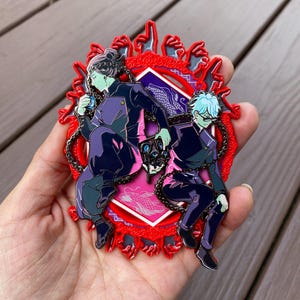 Op de afbeelding: Twee figuren in zwart en paarse kleding zijn aan elkaar geketend en staan op een rode en roze achtergrond met een drakenontwerp. De figuren worden omringd door een rood frame met een ontwerp van handen die naar binnen wijzen.
