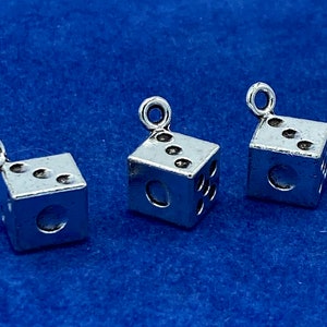 Puede incluir: Tres dijes de dados plateados con un bucle en la parte superior para sujetarlos a un collar o pulsera. Cada dado tiene un número diferente de puntos en cada lado.