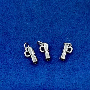 May include: Three silver charms shaped like coffee pots. The charms have a handle and a spout. They are on a blue background.