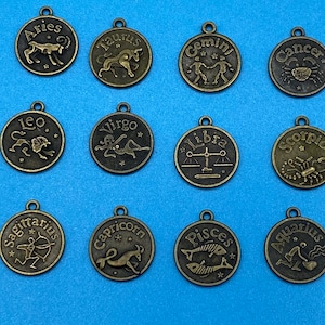 May include: A collection of twelve bronze-colored zodiac sign charms. Each circular charm features a different astrological symbol and the sign's name, including Aries, Taurus, Gemini, Cancer, Leo, Virgo, Libra, Scorpio, Sagittarius, Capricorn, Pisces, and Aquarius.
