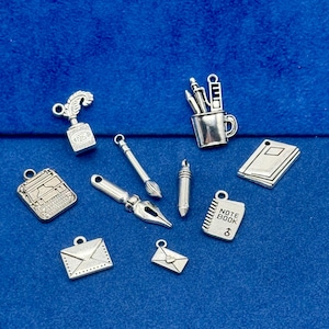 Puede incluir: Una colección de dijes plateados, que incluyen una máquina de escribir, un pincel, un bolígrafo, un bloc de notas y sobres. Los dijes están diseñados con detalles intrincados y probablemente están destinados a la fabricación de joyas o proyectos de artesanía. El fondo es azul liso.